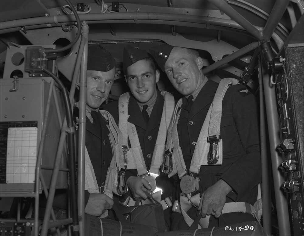 Photographie en noir et blanc – Trois aviateurs tenant des sacs (parachutes?) et portant des sangles se trouvent dans un appareil, entourés d’instruments et de tuyaux en métal. Ils sont tous un peu recroquevillés, car le plafond est bas.